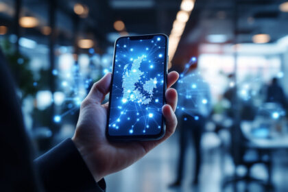 En hånd holder en smartphone, der viser et digitalt kort over Storbritannien med lysende blå netværksforbindelser, i et moderne kontormiljø med slørede mennesker i baggrunden. - Kunstig intelligens AI