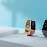 To smartwatches på udstillingsstande: det ene med et farverigt, legende bånd og det andet med et slankt gråt bånd, der begge viser tid og fitnessdata på deres skærme mod en lyseblå baggrund. - Kunstig intelligens AI
